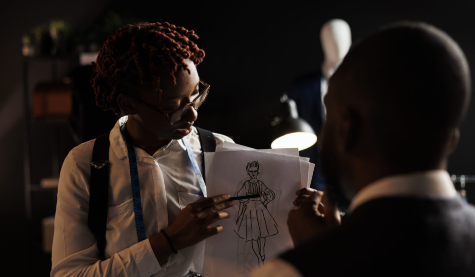 Experienced fashion designer showing couturier sketch drawing of wedding dress she wants him to customize. African american dressmakers working on stylish bespoke sartorial attire